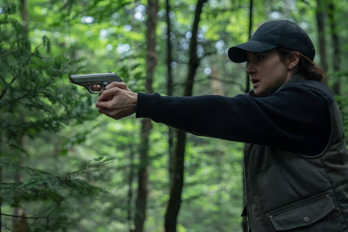 A person wearing a black cap and jacket holds a handgun and aims it in a dense, green forest, appearing alert and focused in a tense outdoor setting.