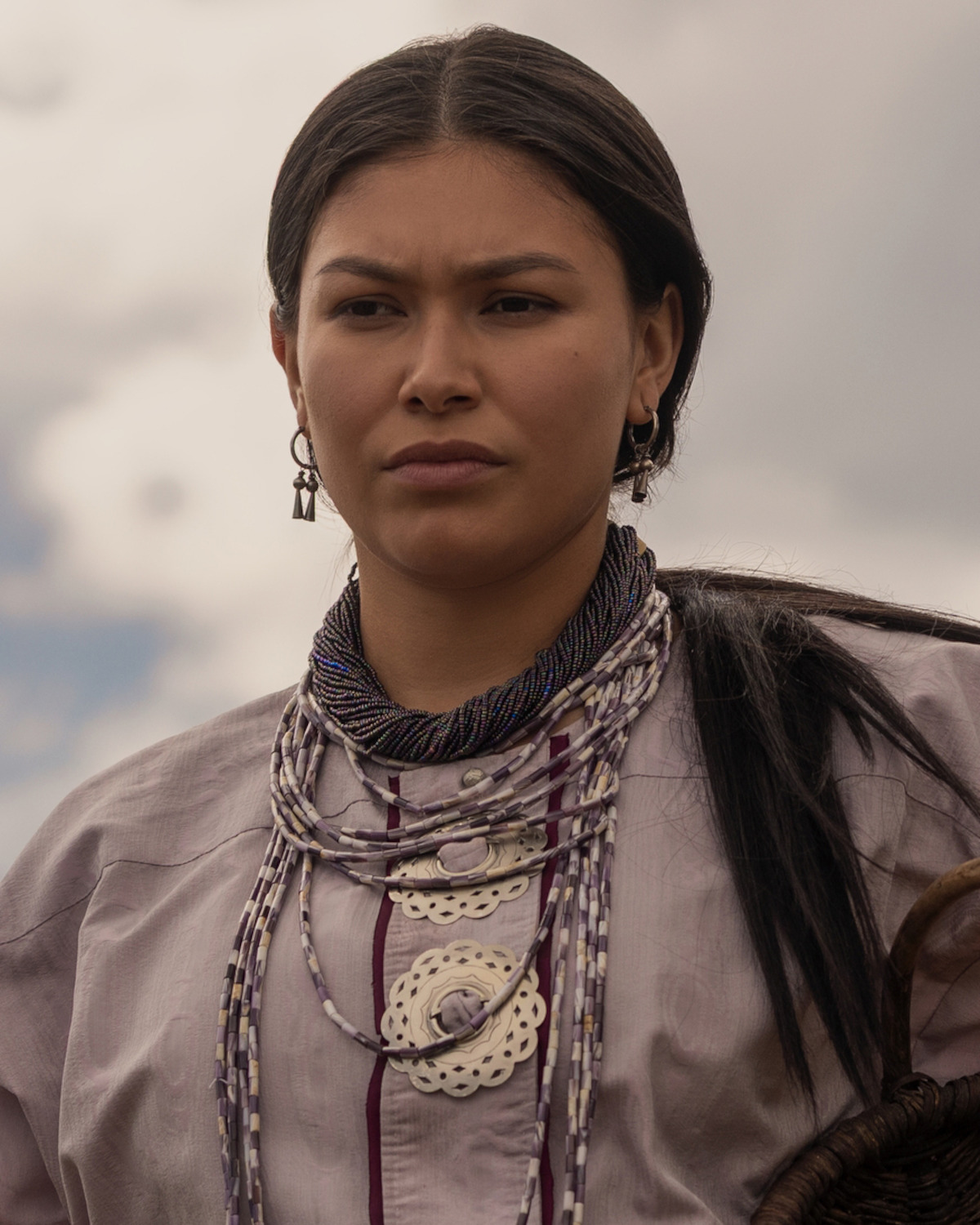 Young woman wearing traditional attire and layered beaded jewelry, standing outdoors with cloudy sky in background, looking serious and composed.