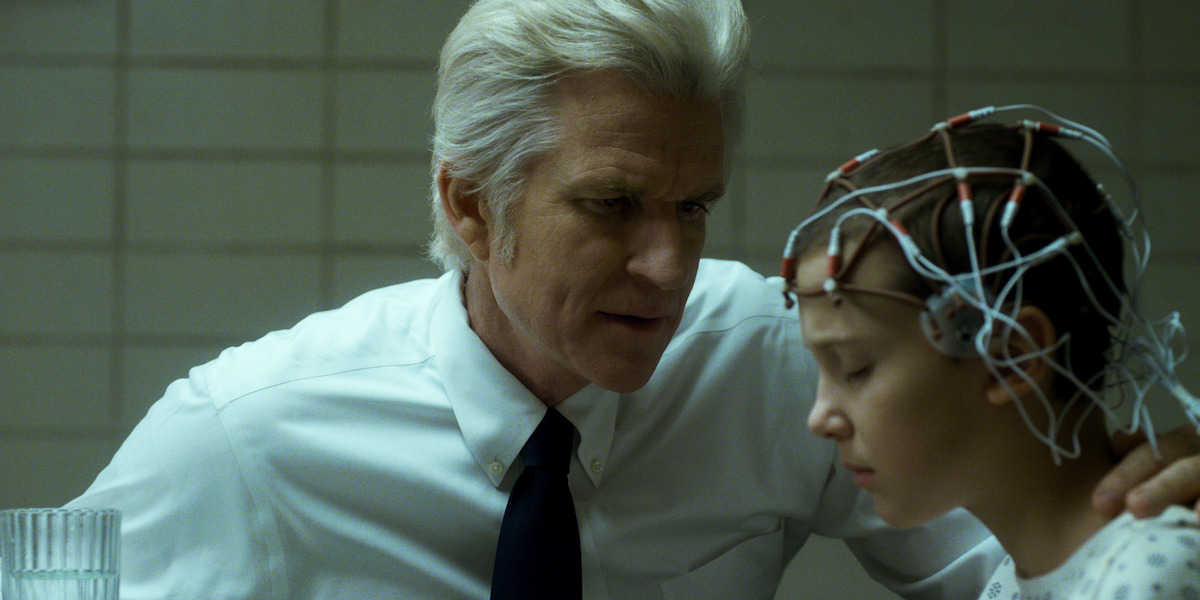 An older white-haired man, Dr. Brenner, sitting next to Eleven who’s wearing a wire harness on her head