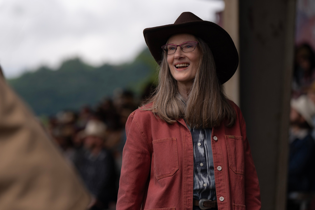 Smiling woman with long hair, pink glasses, and a wide-brimmed hat wearing a red jacket stands outdoors at a western-themed event with blurred people and trees in the background.