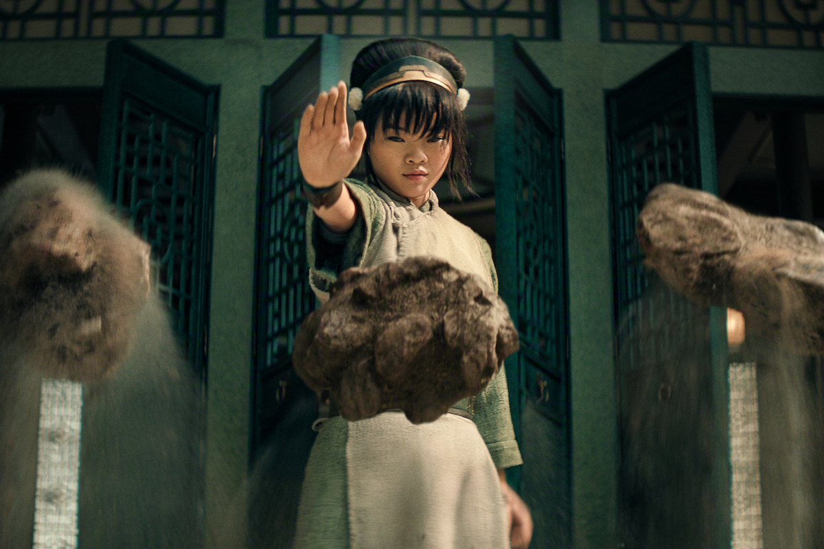 Miyako as Toph in traditional clothing uses a mystical gesture to levitate rocks indoors, with green decorative doors in the background and sand trickling from the floating stones.