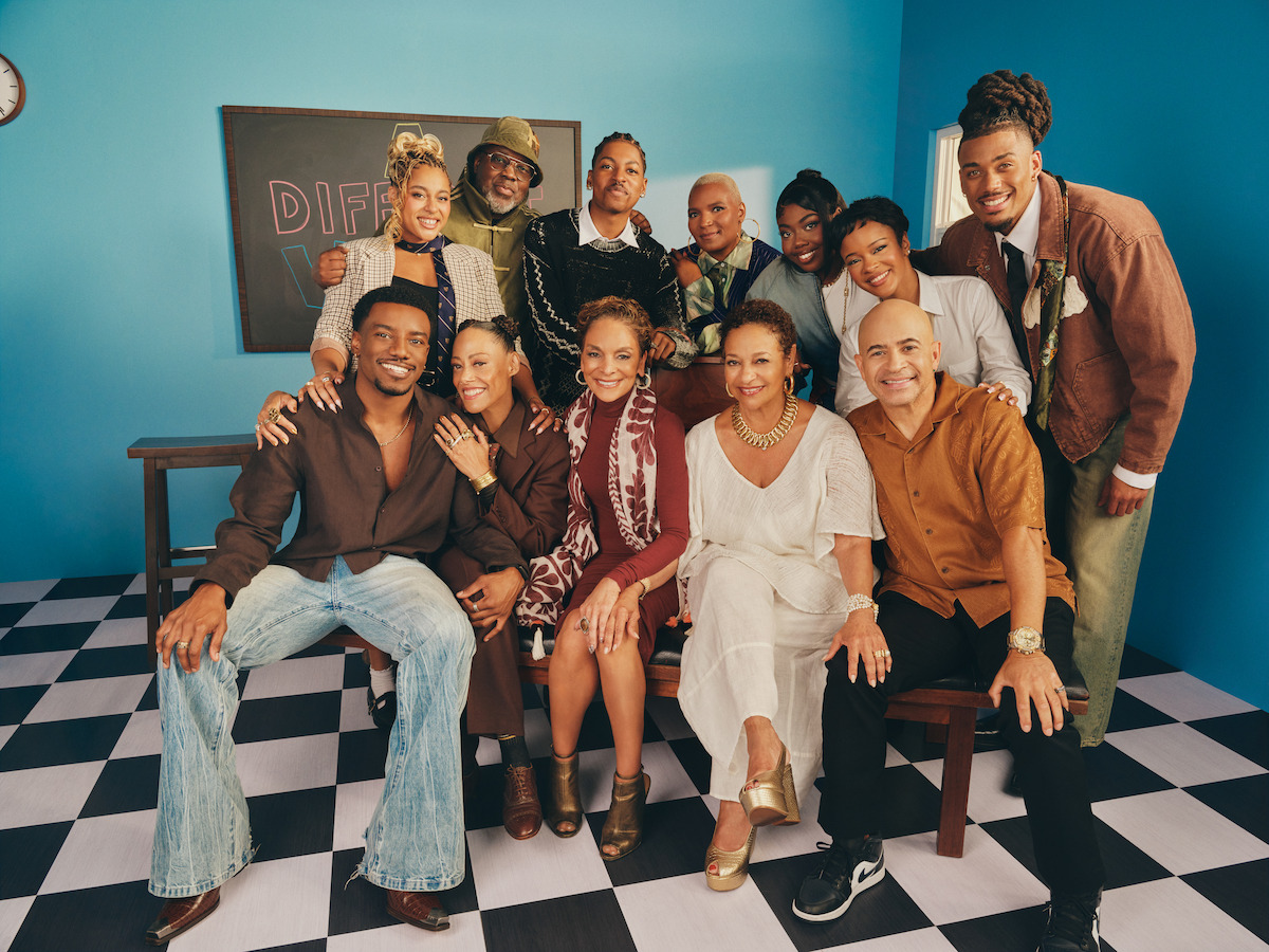 Group of people posing and smiling together in a bright, retro-themed room with a blue wall and black-and-white checkered floor, bench seating, chalkboard, and vintage decor visible in the background.