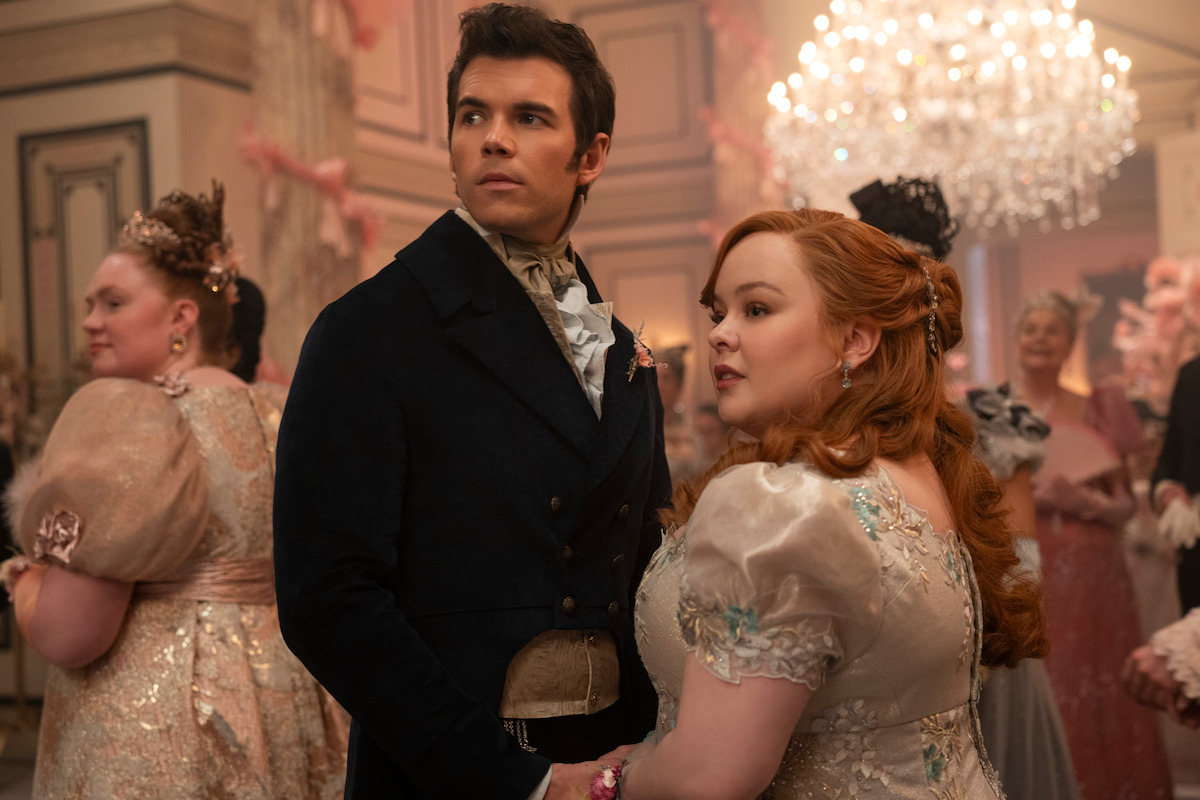A man and woman in period costumes stand together at an elegant ballroom event, surrounded by other guests in ornate dresses with a grand chandelier and lavish decor in the background.