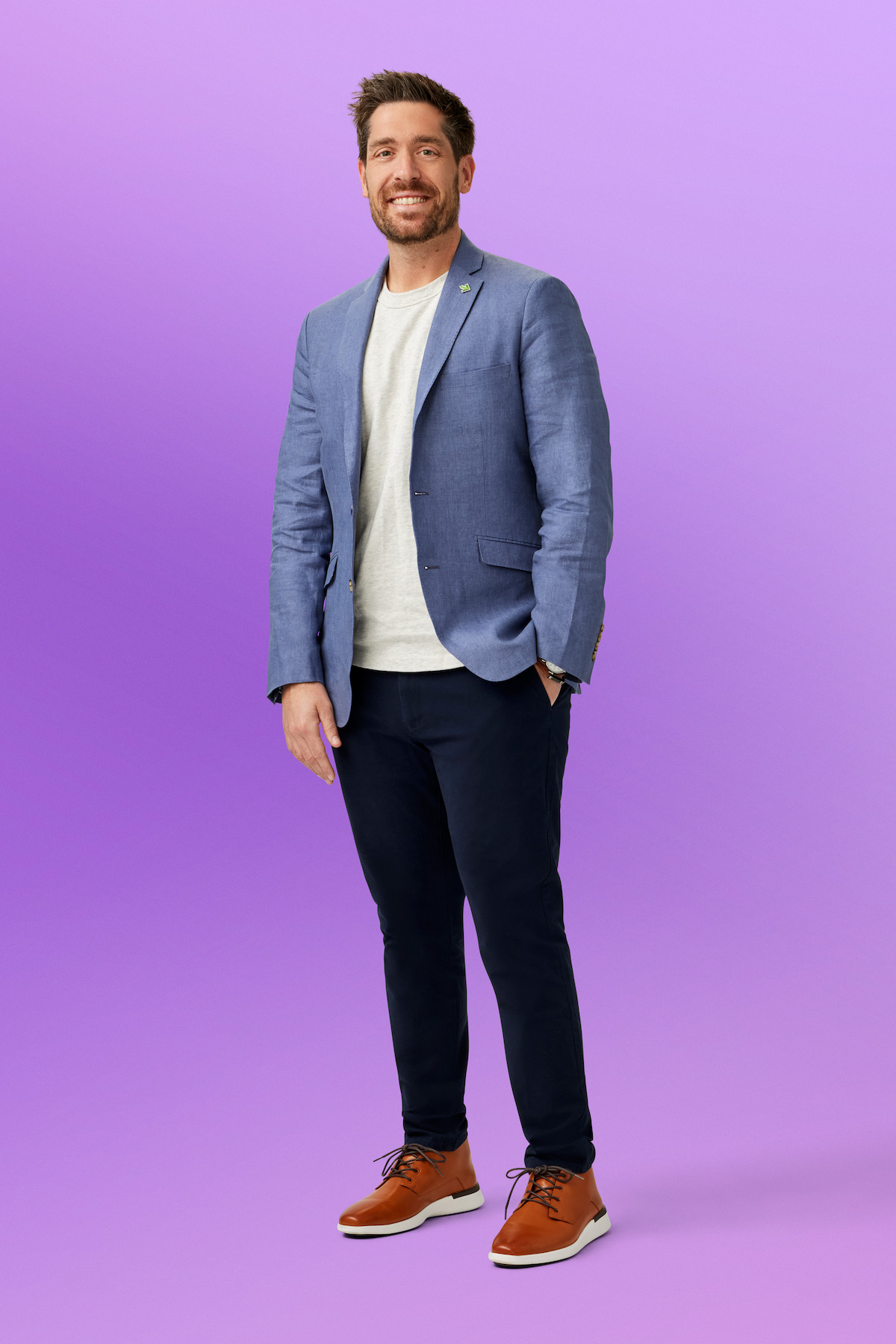 Kevin smiling, wearing a blue blazer, white shirt, dark pants, and brown shoes in a studio with a light purple background.