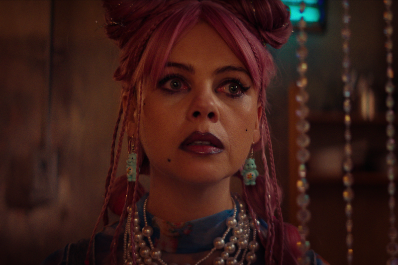 Woman with pink hair styled in buns, dramatic makeup, and layered jewelry stands indoors with warm lighting and beaded curtain in background, looking serious or contemplative.