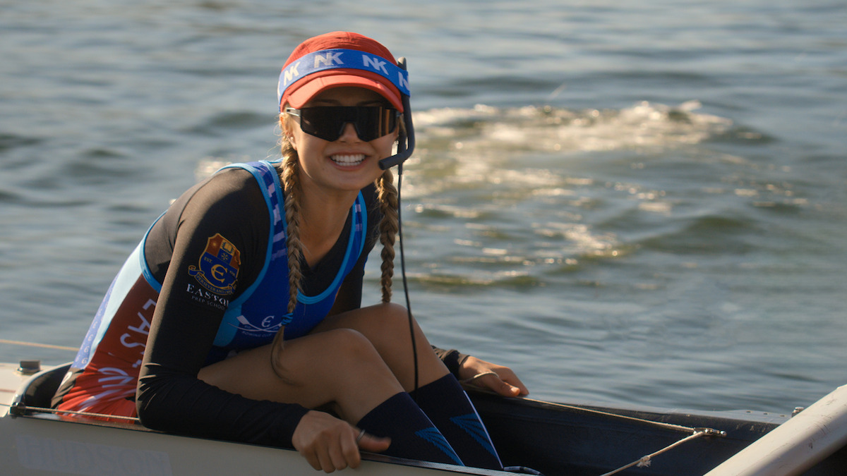 Smiling female rowing coxswain in a boat on the water, wearing sunglasses, a headset, and a team uniform with a red visor, during a sunny day.