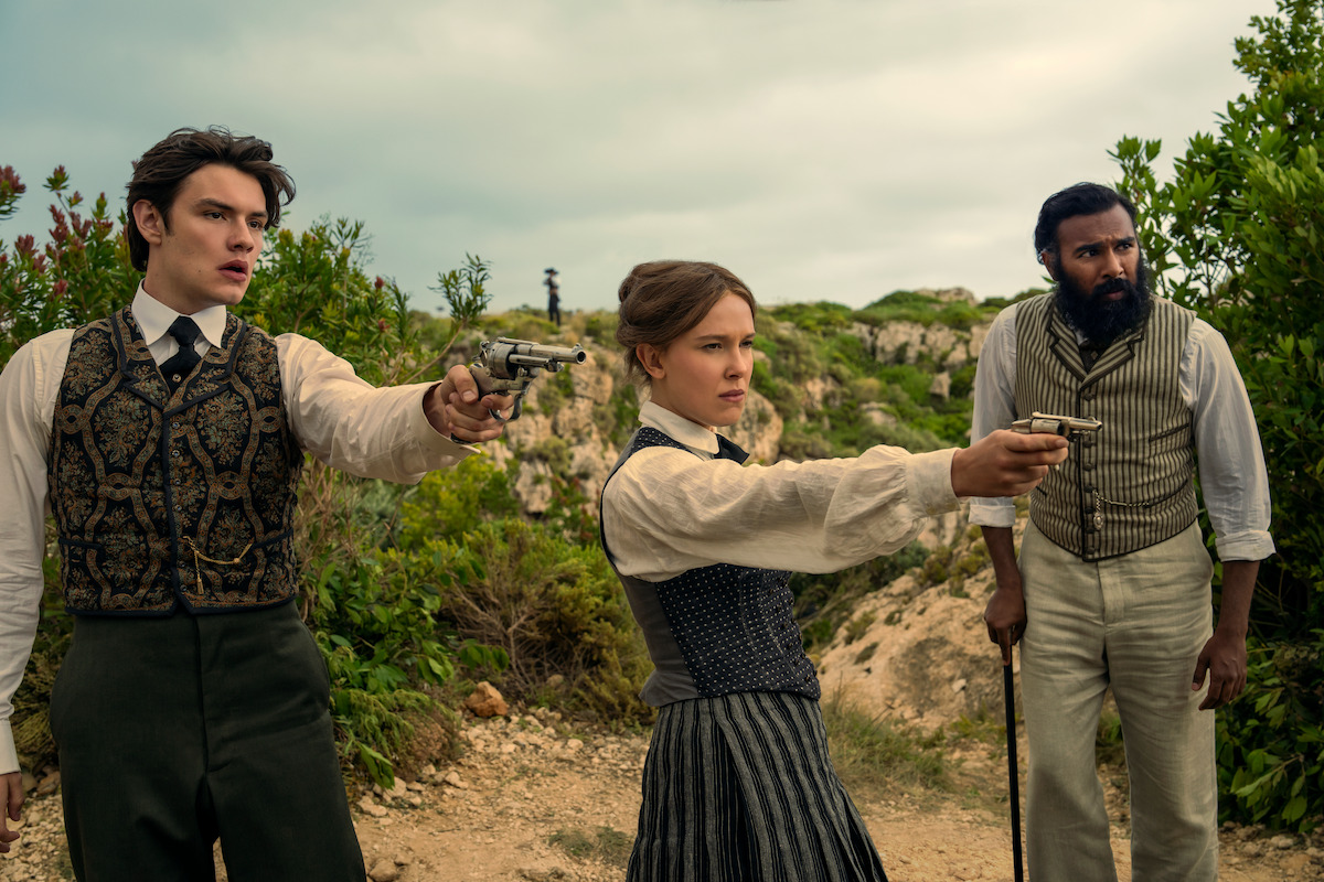 Three people in vintage clothing stand outdoors in a rugged, green landscape; two point pistols ahead while the third stands beside them with a cane, all looking alert and tense.
