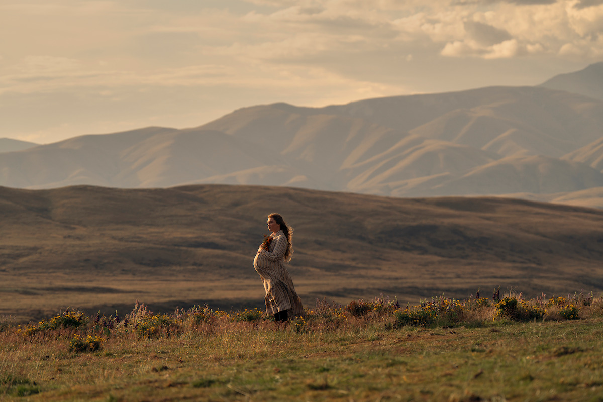Pregnant woman standing in a grassy field at sunset, surrounded by wildflowers, with rolling hills and mountains in the background under a cloudy sky.