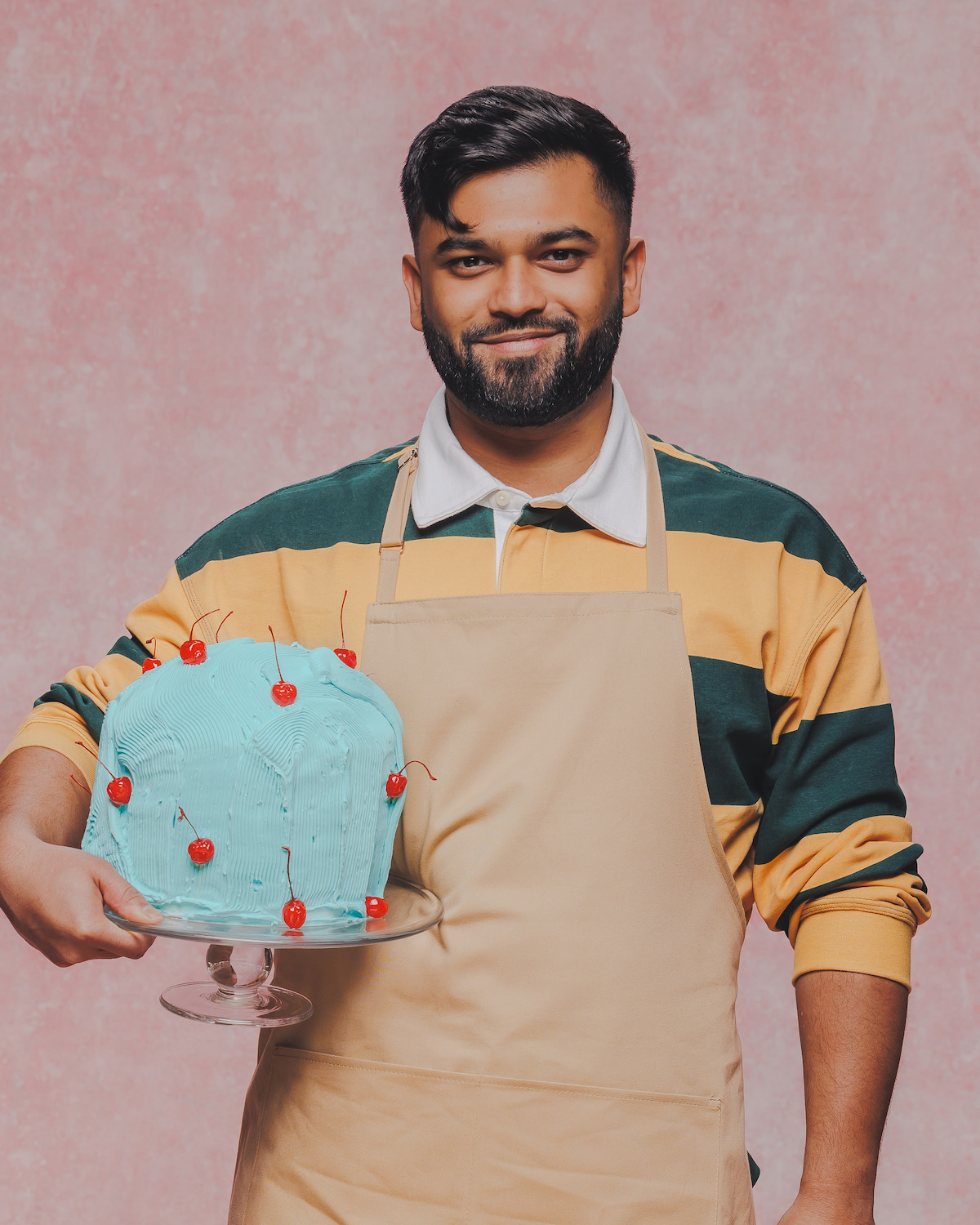 Man wearing a tan apron and striped shirt holds a blue frosted cake with cherries on a glass stand, standing in front of a pink textured background.