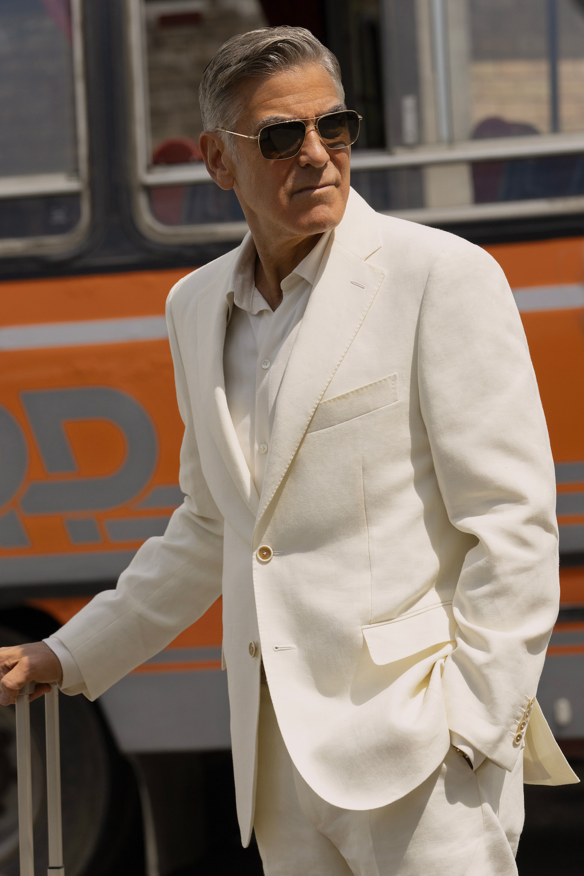 Man in a white suit and sunglasses stands confidently outdoors near an old-style bus, holding a suitcase, with a relaxed yet stylish mood.