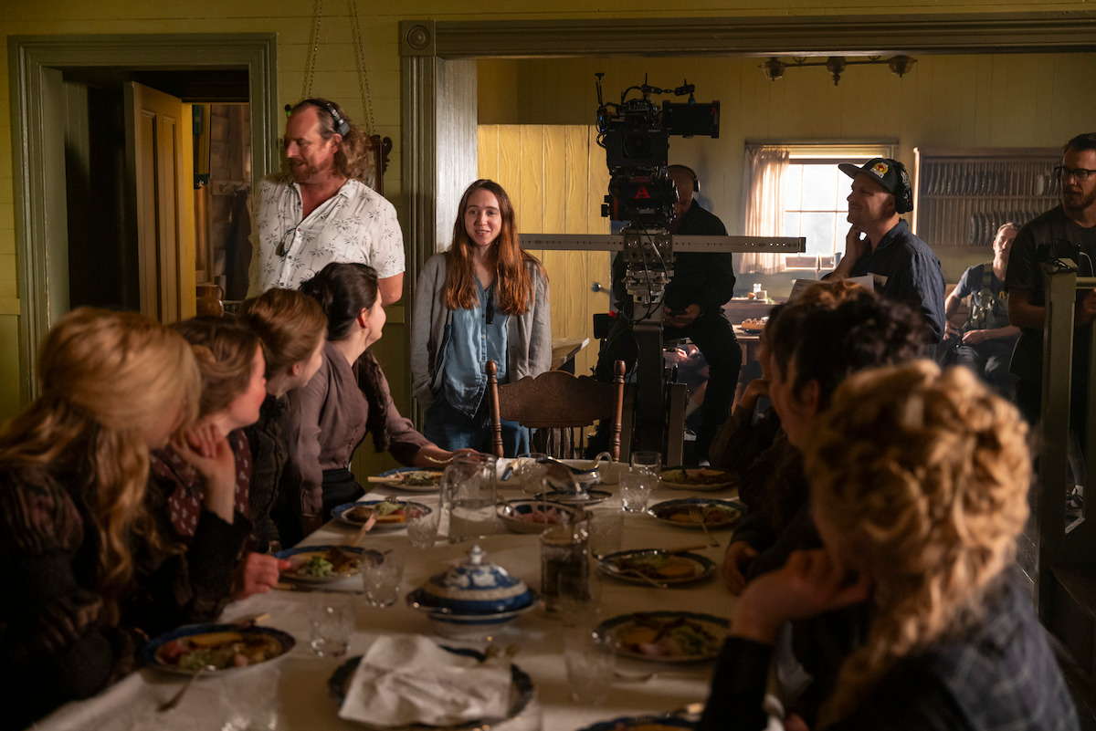 Film crew and actors gather around a dining table set with food on a film set, with cameras and lights visible, capturing a period drama scene in a warmly lit room.