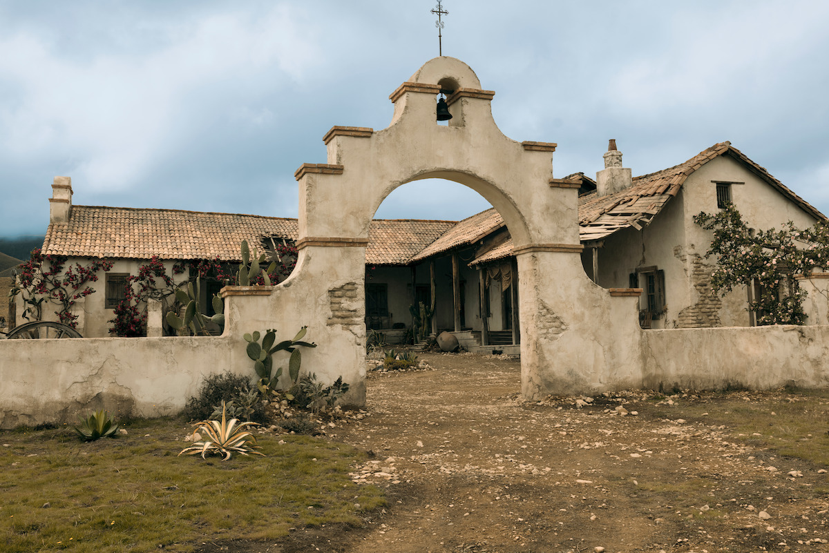 Old adobe-style house with arched entry gate, weathered walls, tile roof, and surrounding desert landscape with cacti under cloudy sky.