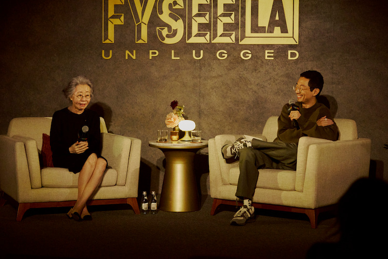 Two people sit in armchairs on a stage talking into microphones at a "FYSEE LA Unplugged" event, with a small table between them holding drinks and flowers, against a warmly lit backdrop.