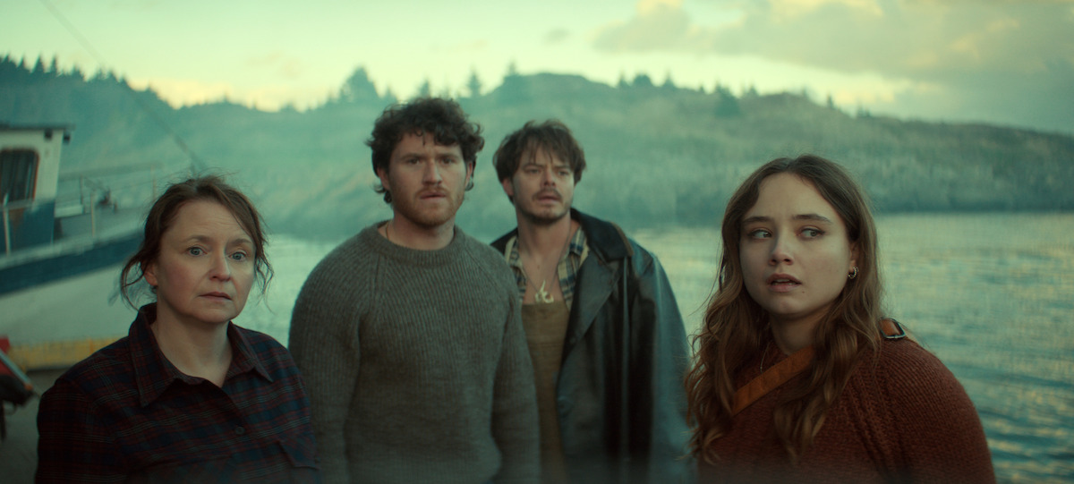 Four people stand near a waterfront, looking concerned. Behind them is a boat and a rocky shoreline with pine trees under a cloudy sky. The lighting is moody, suggesting a dramatic or tense atmosphere.