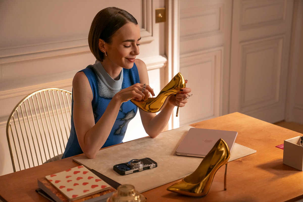 Woman sitting at a desk in a stylish, softly lit room, smiling and examining a shiny gold high-heeled shoe. The desk has notebooks, a phone, a laptop, and another gold shoe, creating a creative work environment.