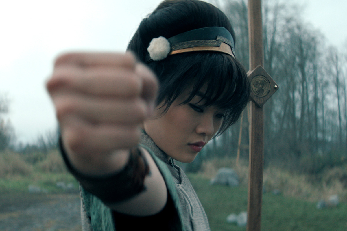 Miyako as Toph with short dark hair, dressed in fantasy-style clothing, poses with her fist forward in a fighting stance outdoors in a misty field with trees and a staff or spear visible behind her.