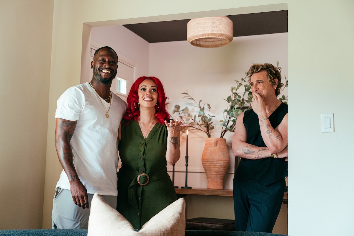 Three people stand and pose in a cozy, modern living room. Decorative vases, plants, and soft lighting create a relaxed, inviting atmosphere. One person smiles, another gestures, and the third stands thoughtfully near a doorway.