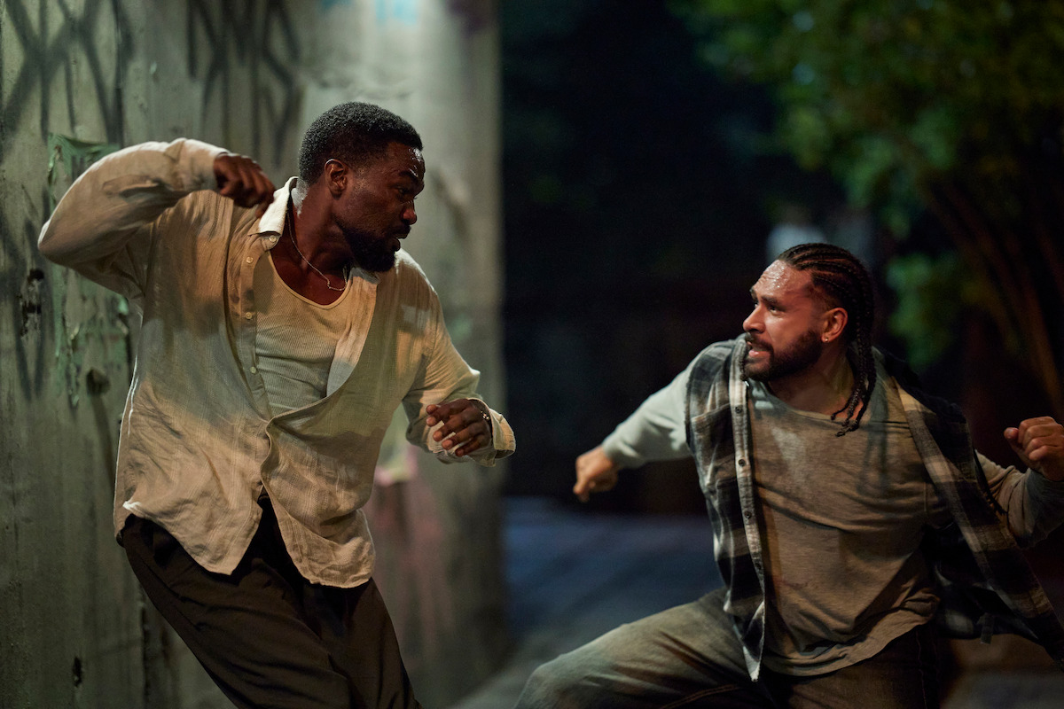 Two men are fighting in a dimly lit urban alley at night, with tense expressions and raised fists, surrounded by graffiti-covered walls and trees in the background.