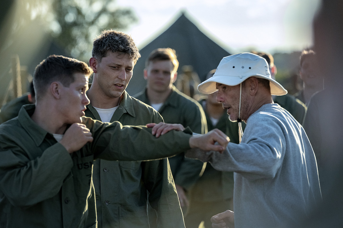 Military recruits practice hand-to-hand combat outdoors, wearing green uniforms, with a trainer in a bucket hat instructing them; tents and trees visible in the background, group setting, training camp atmosphere.