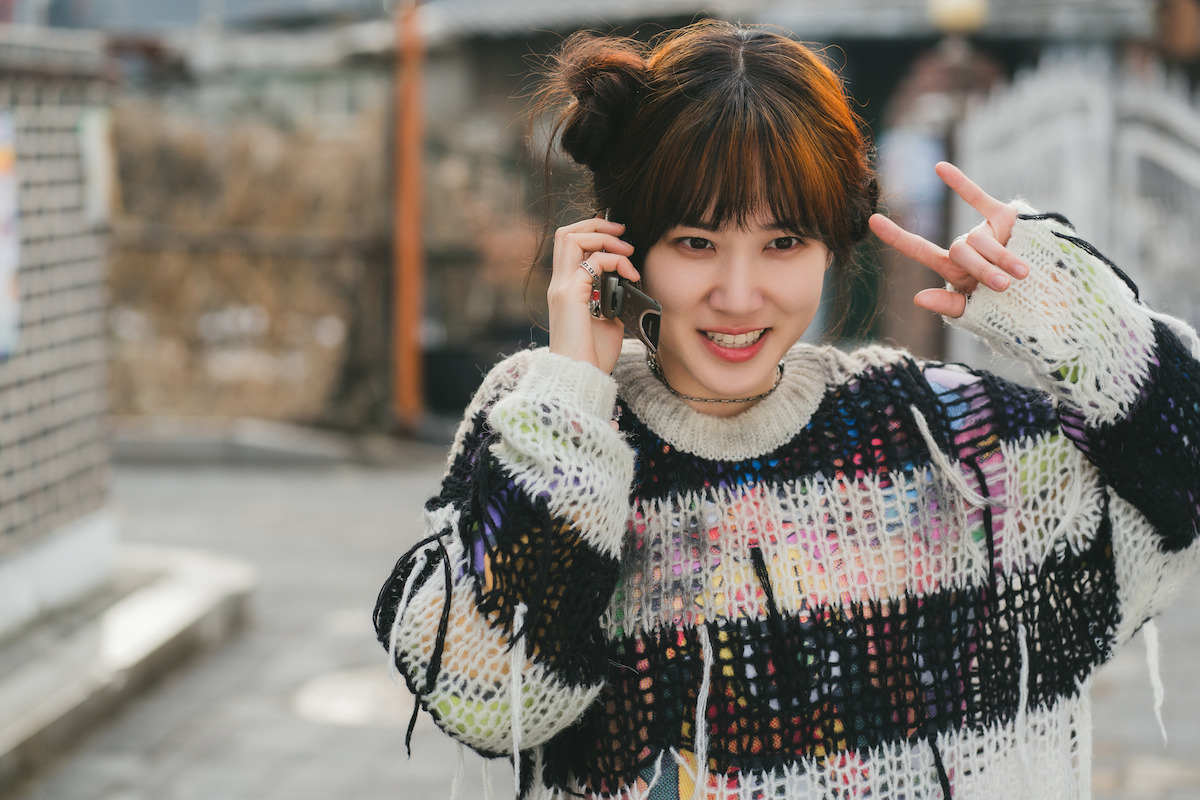 Young woman outdoors wearing a colorful sweater, smiling while talking on the phone and making a peace sign with her hand, blurred urban street in the background.