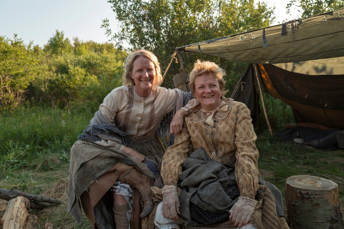 Two women in pioneer-era clothing sit smiling at a rustic campsite with a canvas tent behind them, surrounded by greenery and sunlight, evoking a historical outdoor setting.