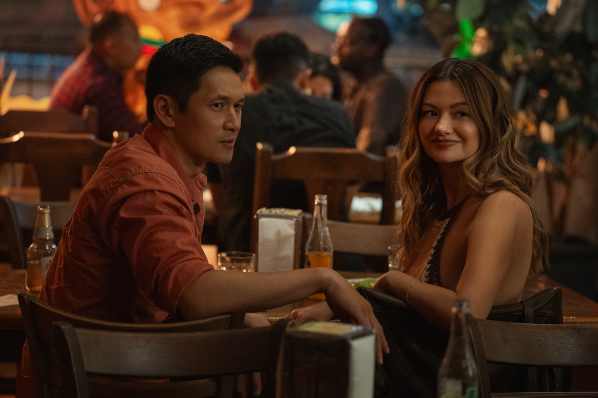 A man and woman sit together at a wooden table in a dimly lit, busy restaurant or bar, with drinks and napkins on the table, surrounded by other patrons in the background.