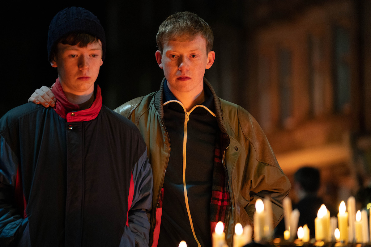 Two young men stand together at a nighttime candlelight vigil, looking somber. One has his arm around the other for comfort. The setting is outdoors in an urban area, with lit candles in the foreground.