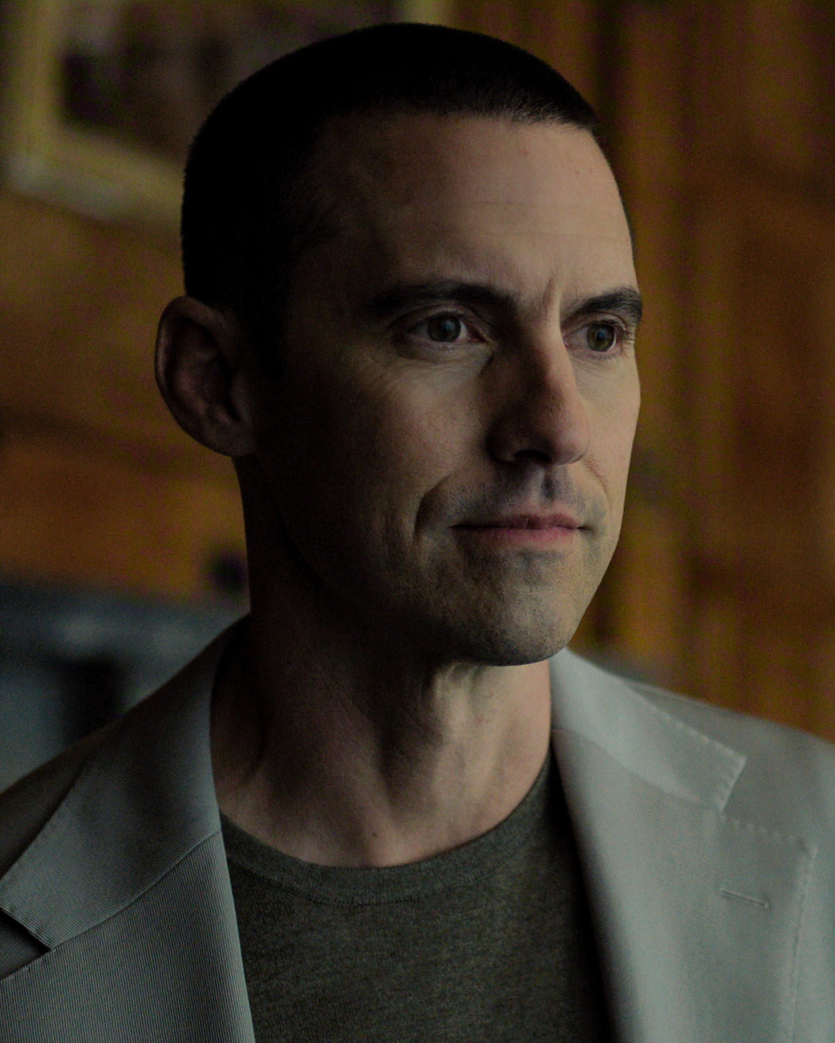 A man with short dark hair wearing a light-colored blazer and t-shirt stands indoors in a warmly lit room with wood-paneled walls and a soft, focused expression.