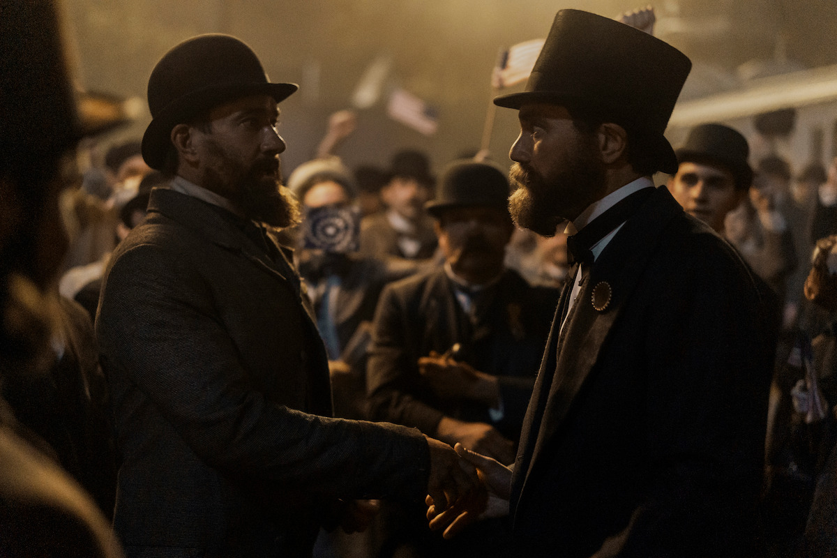 Two men in 19th-century attire shake hands in a dim, crowded outdoor setting, surrounded by onlookers with hats and flags, creating a formal, historical, and tense atmosphere.
