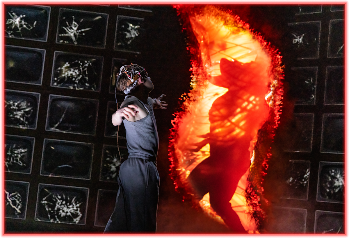 A person in a futuristic outfit with headgear faces a fiery portal showing a dramatic silhouette, set against a wall of dark, abstract screens with a glowing red border.