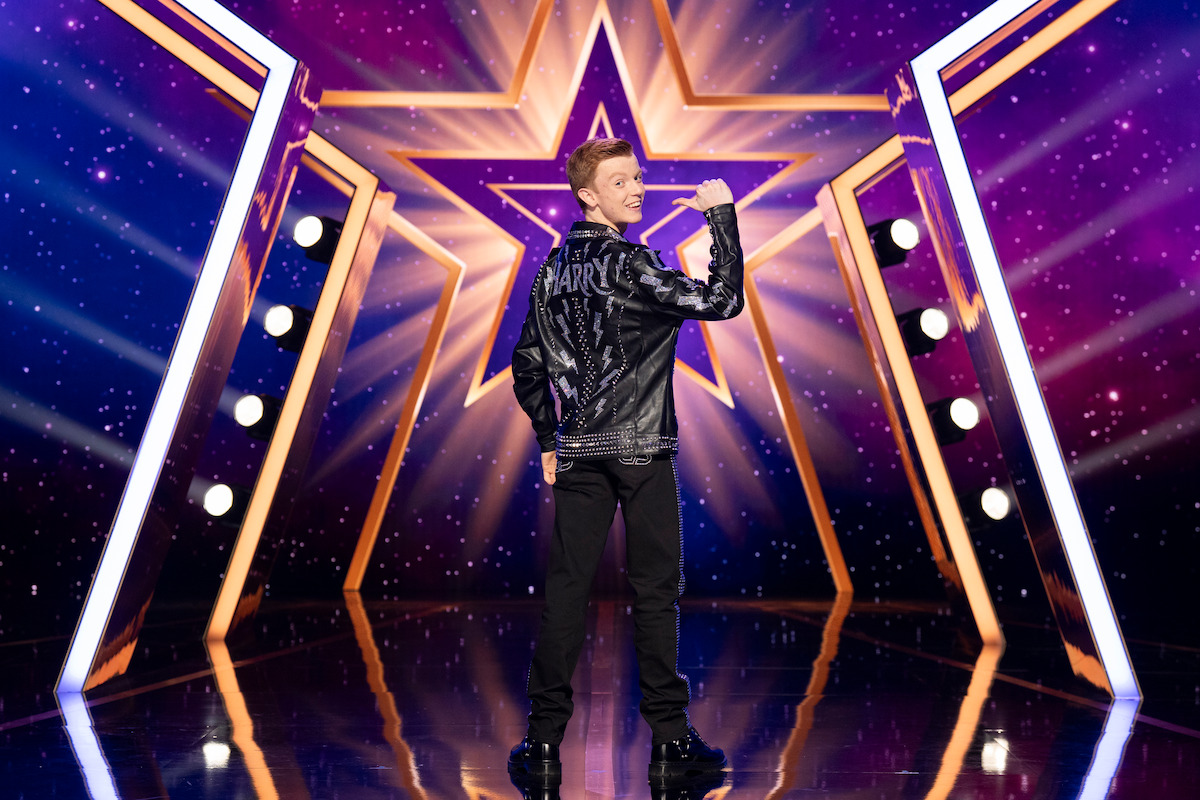 Harry Merlin Piper stands on a shiny stage with a large illuminated star and spotlights, facing away and looking over their shoulder, in a vibrant, colorful show environment.