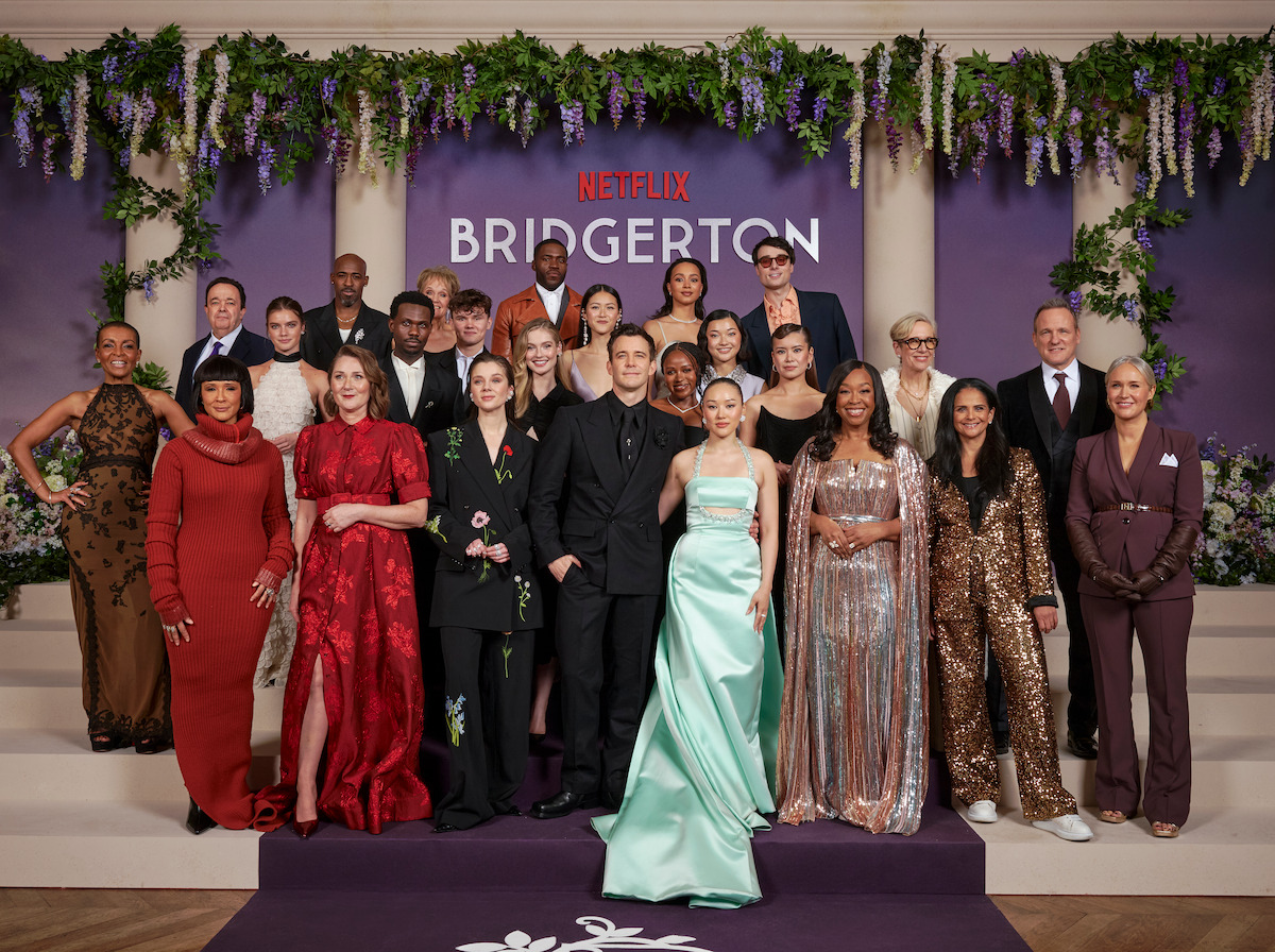 A group of elegantly dressed people posing on steps at a formal Netflix Bridgerton event, with floral decorations, purple backdrop, and lush greenery in the background.