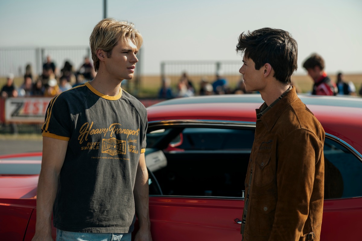 Two young men are standing beside a red car, facing each other seriously in an outdoor setting with a group of people and a fence in the background, possibly at a race event or car meet.