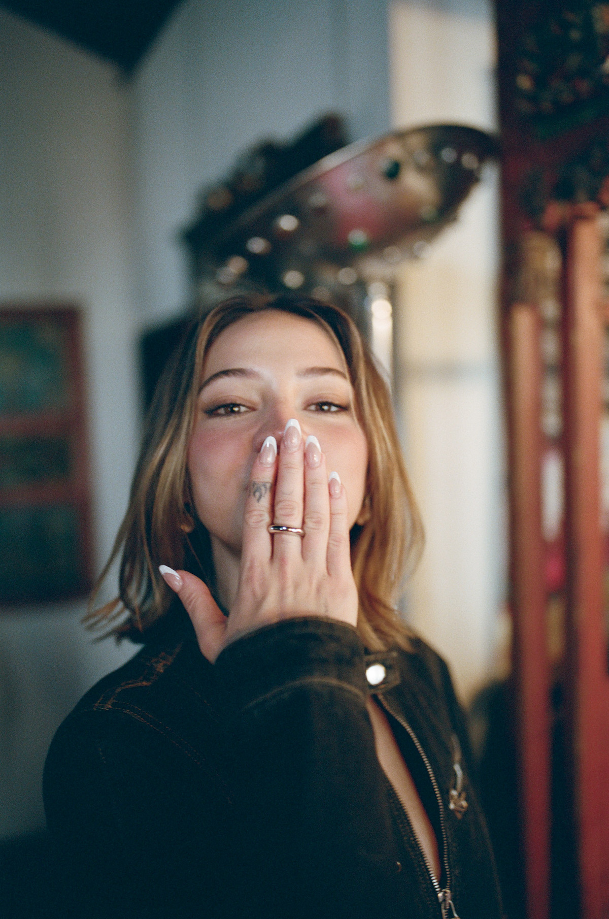 Woman with light brown hair blowing a kiss indoors, wearing a dark jacket. The background is softly focused, with warm lighting and decorative elements, giving the scene a cozy, inviting atmosphere.
