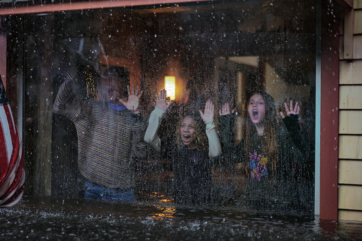 Three people inside a house stand behind a large window, pressing their hands and faces to the glass, looking distressed as heavy rain and flooding occur outside. The room is dimly lit and water rises against the window.