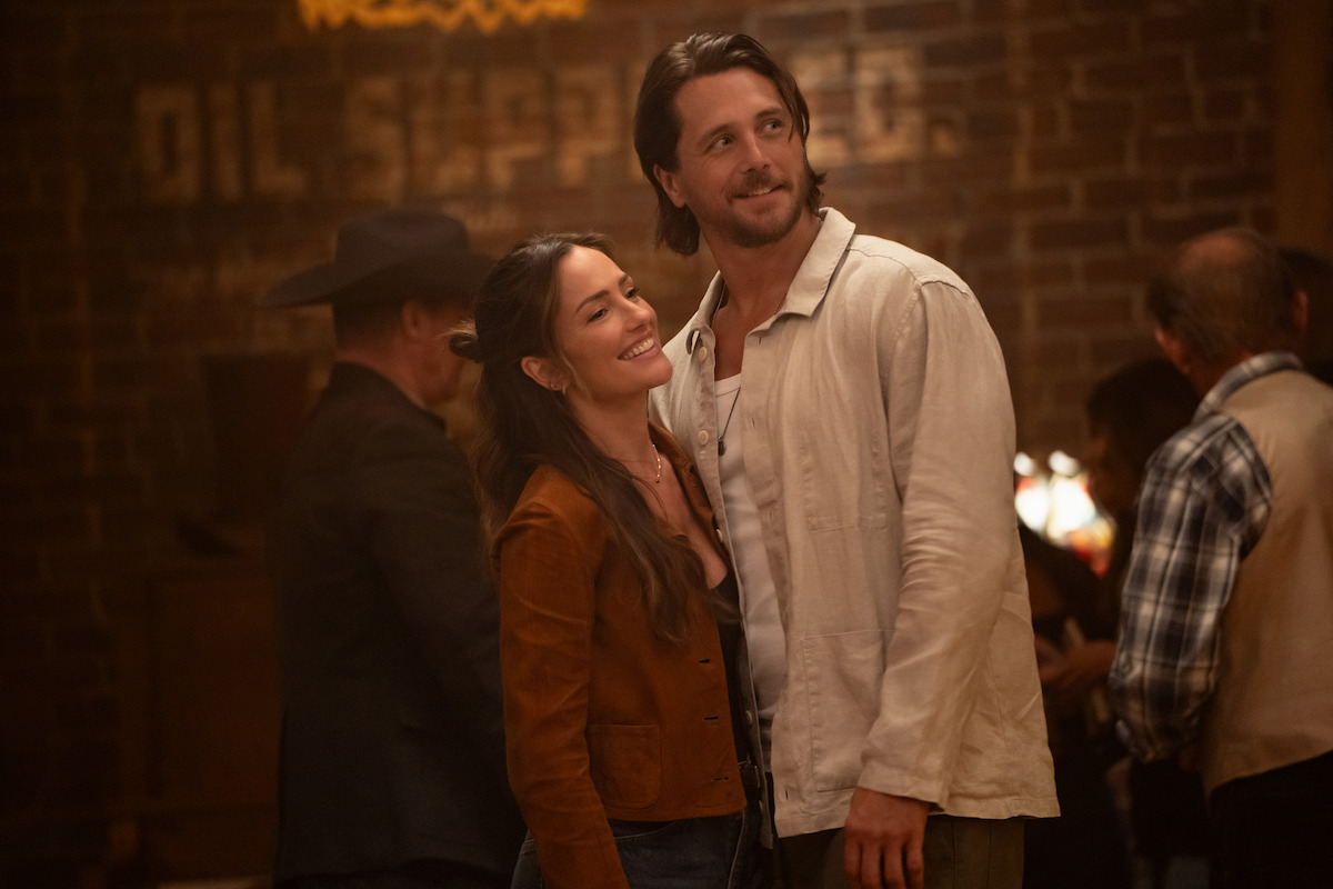 A smiling couple stands together in a warmly lit rustic indoor setting, surrounded by people socializing. The background features exposed brick walls and a vintage painted sign, creating a cozy, welcoming atmosphere.