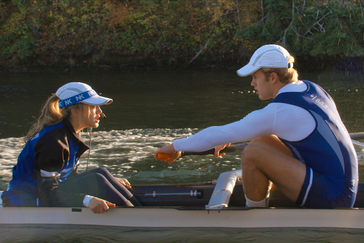 Two rowers in blue uniforms, one male and one female, are in a boat on a river. Trees and reflections are visible in the background.