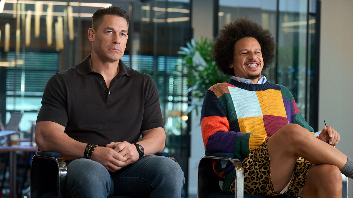 Two men sitting side by side in a modern office setting; one wears a black polo and jeans looking serious, the other wears a colorful sweater and shorts, smiling and relaxed. Large windows and plants are in the background.