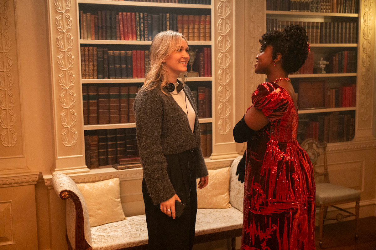 Two women stand and smile at each other in a richly decorated library room, one wearing a red sequin dress and the other in casual attire with headphones around her neck, surrounded by bookshelves and ornate furniture.