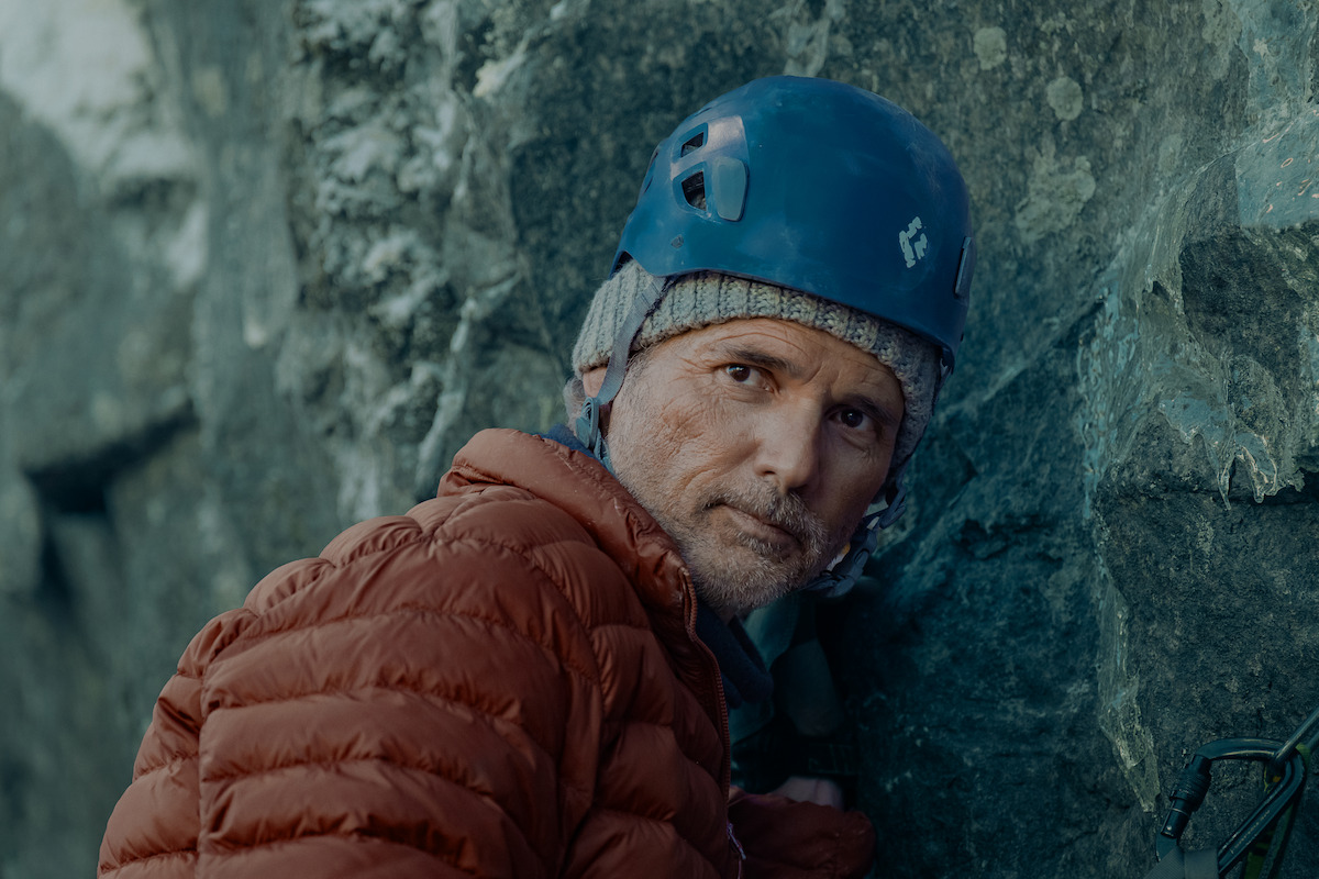 A climber in a blue helmet and red jacket rests against a rocky cliff outdoors, focused and geared for cold weather.