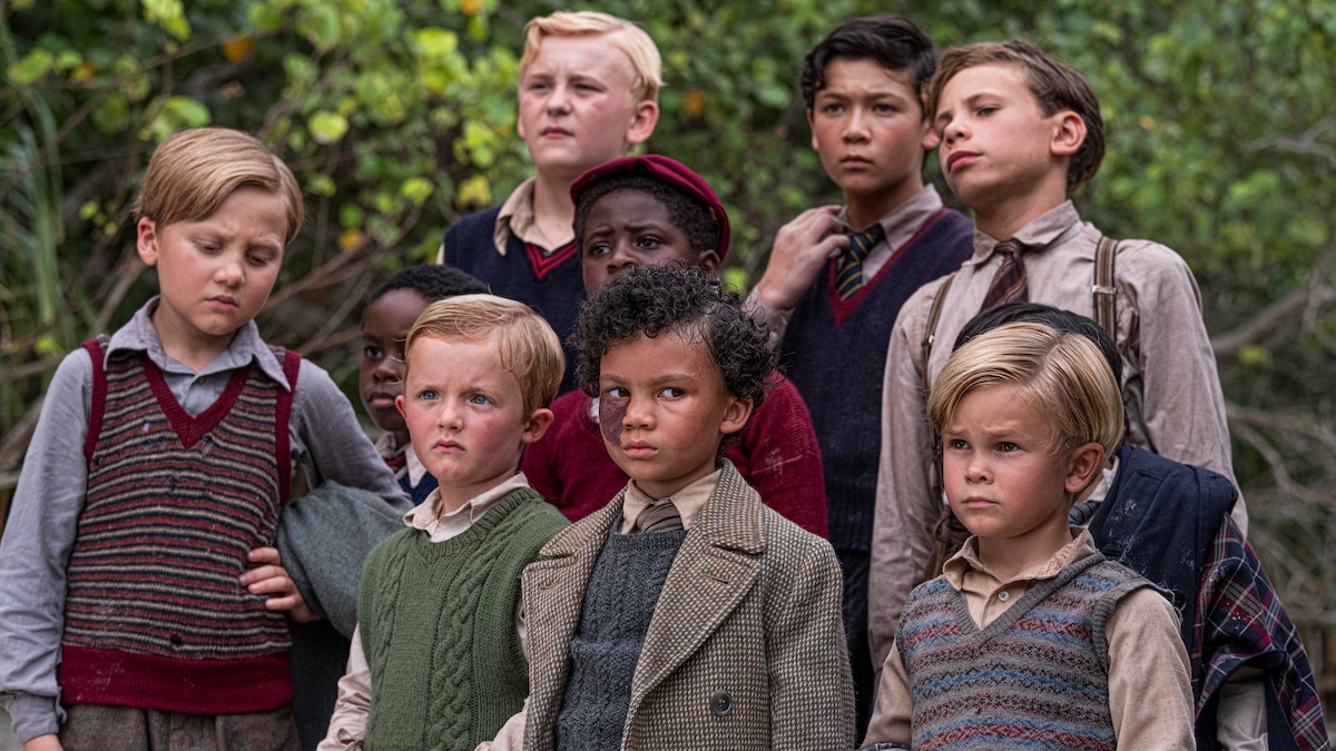 Group of young boys in vintage clothing standing outdoors with trees and greenery behind them, serious expressions, appearing to be in a forest or natural setting.