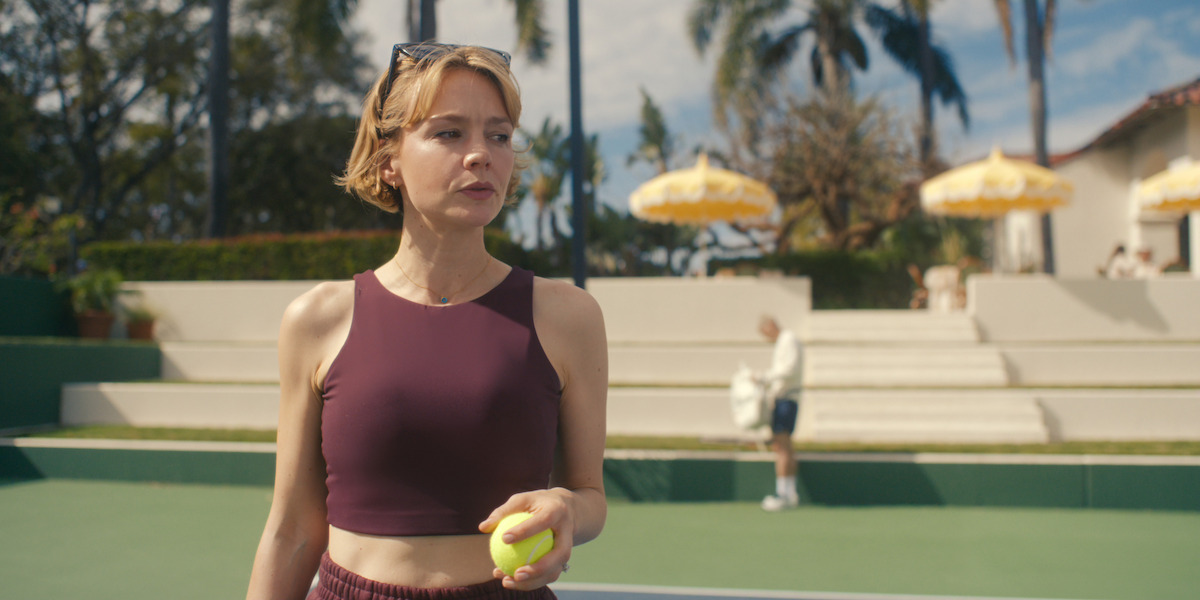 Woman in athletic wear holding a tennis ball on an outdoor tennis court with palm trees, yellow umbrellas, and steps in the background under sunny skies.