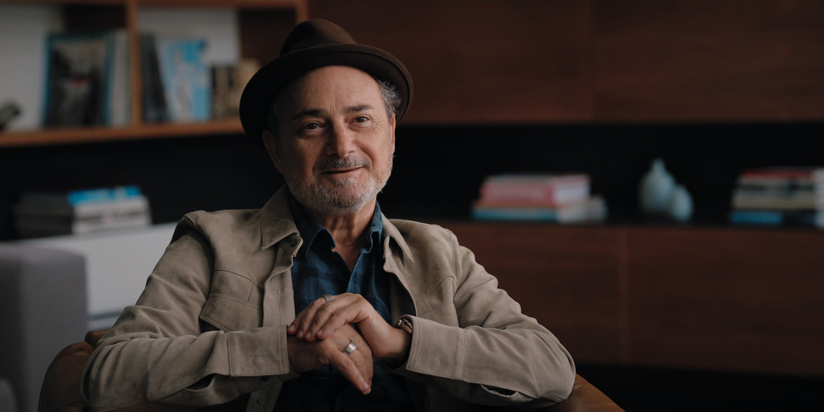 A smiling man in a brown hat and beige jacket sits indoors with bookshelves and a modern background, creating a relaxed, intellectual atmosphere.