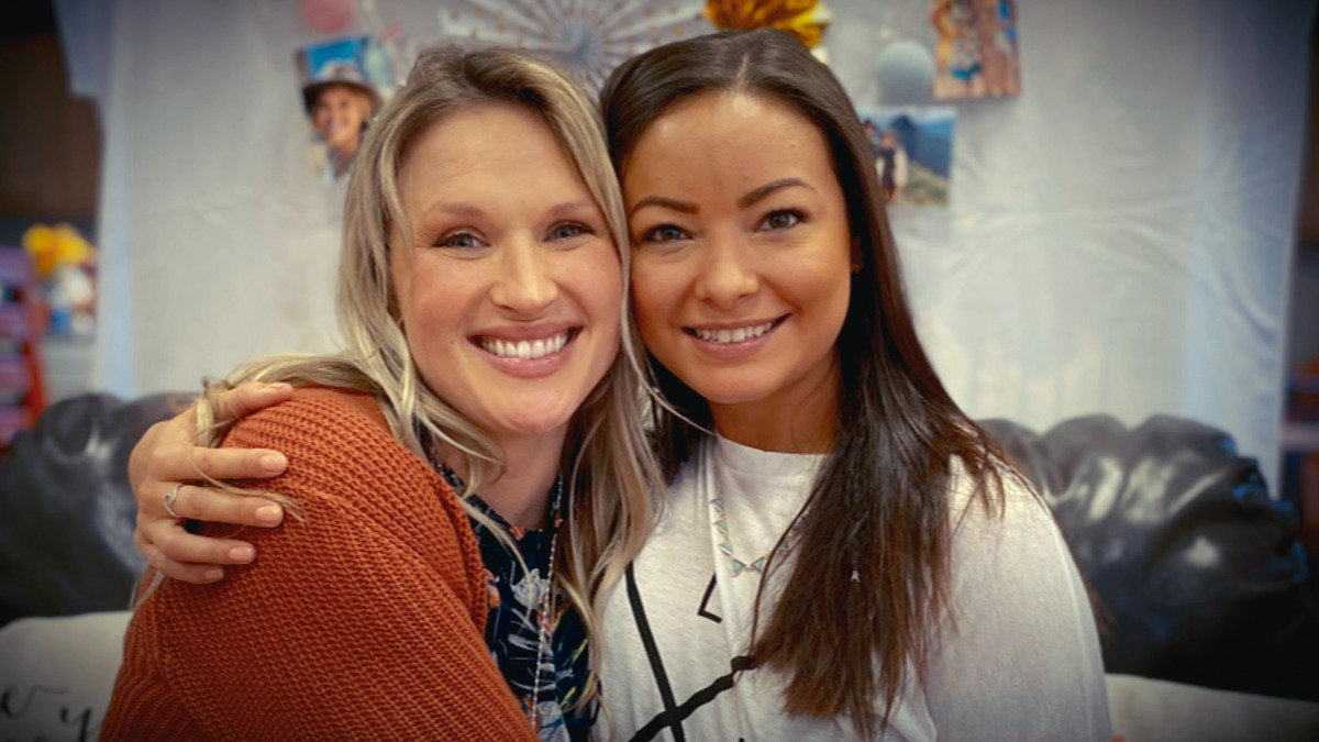 Two women smiling and sitting close together on a black couch, embracing each other in a cozy indoor setting with photos and decorations in the background.