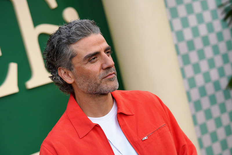 A man with wavy gray hair and a beard wearing a bright red jacket over a white shirt stands outdoors in front of a green and white checkered backdrop, looking off to the side.