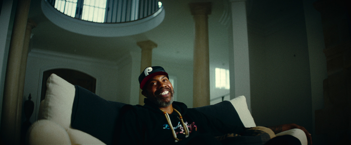 Rasheed Wallace, con una gorra de béisbol y una sudadera con capucha, está sentado sonriendo en un sofá en una sala de estar espaciosa y exclusiva con columnas altas, ventanas grandes y una suave luz natural que se filtra.