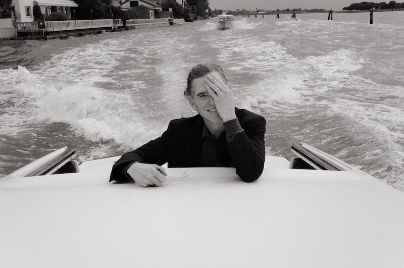 Billy Crudup covers half his face with his hand while standing on a boat with the Venice canals behind him.