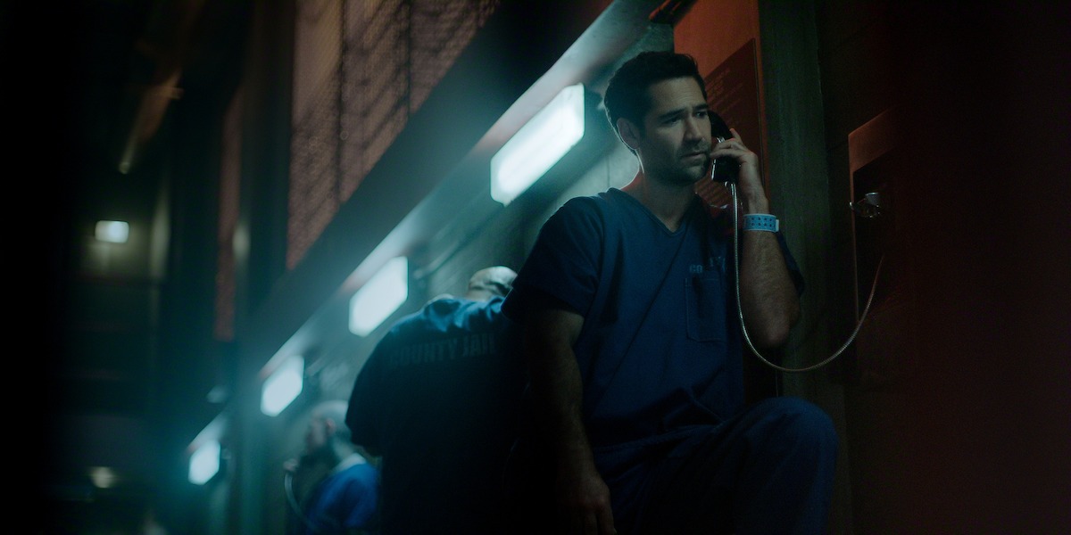 A man in blue jail scrubs sits in a dimly lit jail visiting area, using a wall-mounted phone, with fluorescent lights above and other inmates in the background making calls.