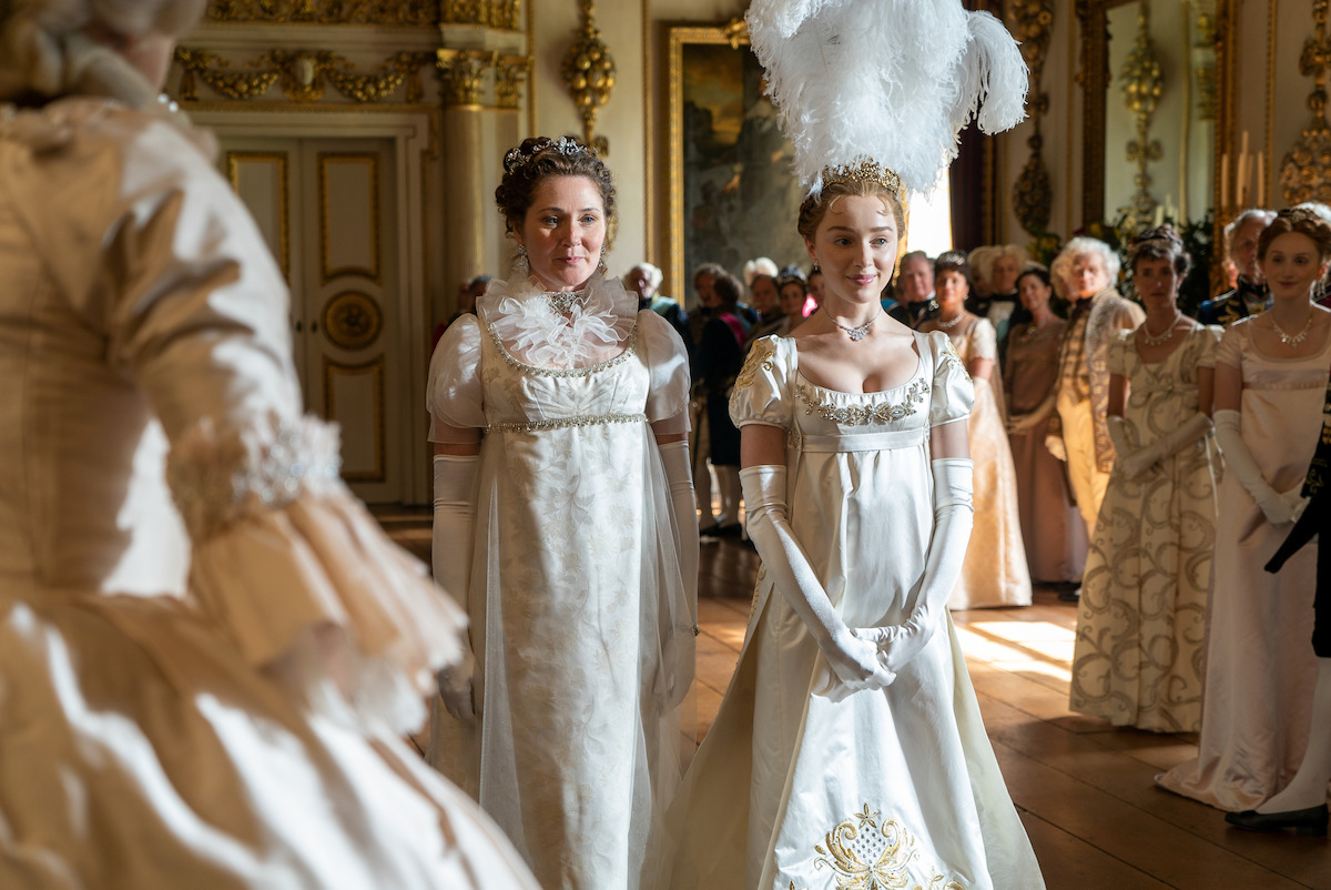 Two women in elegant white Regency-era gowns and gloves stand in a grand, ornate ballroom with gold accents, crowds of guests in period attire, and sunlight streaming through large windows, evoking a historical or royal event atmosphere.