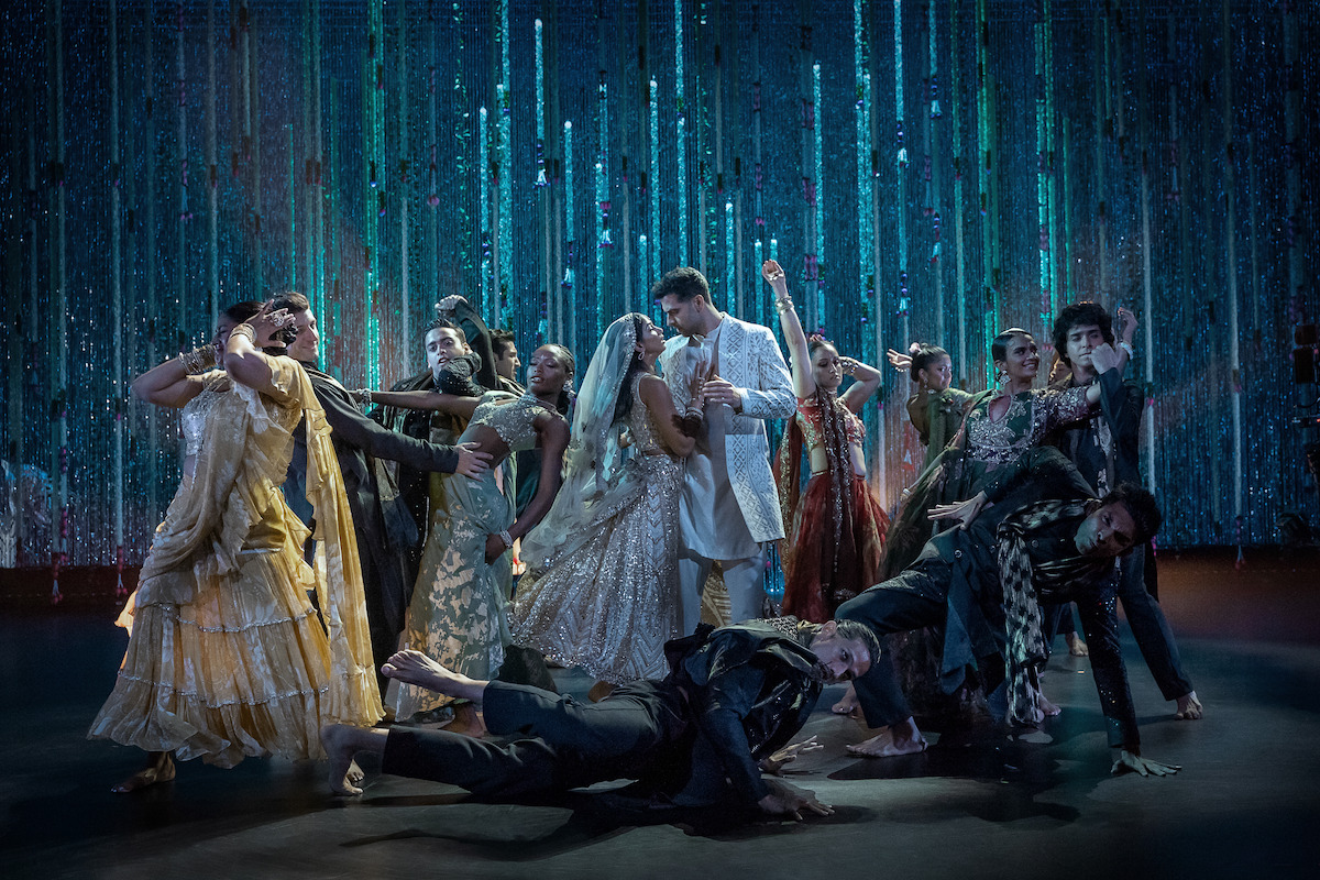 A group of dancers in elaborate, sparkling outfits perform a dramatic scene on stage with a glittering blue curtain backdrop, evoking a festive or celebratory atmosphere.